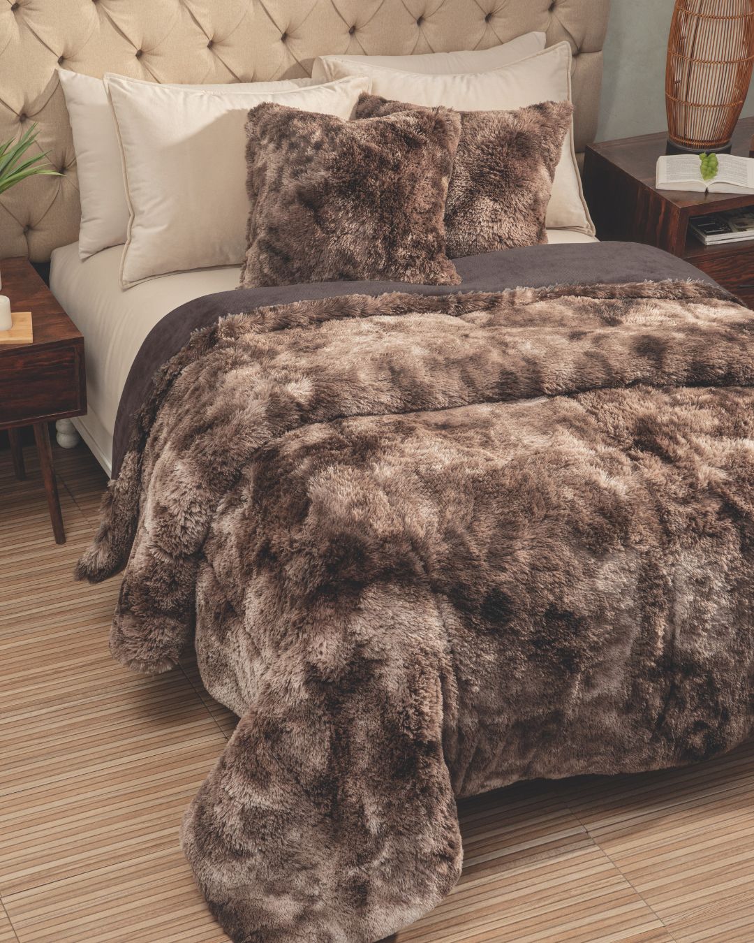 A bed with a beige tufted headboard is neatly made with the Intima Hogar Cobertor Platinum Extra Suave Montana, complemented by matching pillows and white accent pillows. Wooden nightstands on each side display decorative items.