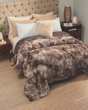A cozy bedroom with a tufted headboard, neatly arranged pillows, and the Intima Hogar Cobertor Platinum Extra Suave Montana—a fluffy brown faux fur blanket—spread on the bed, flanked by two wooden nightstands with lamps, a plant, and a small framed photo on one side, all set atop a wooden floor.