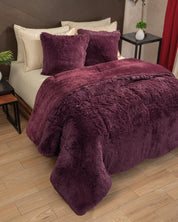 A neatly made bed features the Cobertor Platinum Extra Suave Nevada by Intima Hogar in a rich, deep purple, paired with matching pillows. The modern room is accented by wooden floors, cream pillowcases, a dark headboard, and tasteful decor on the bedside tables. The textura ultra suave of this bedding brings an added layer of calidez, making the space feel exceptionally inviting and comfortable.