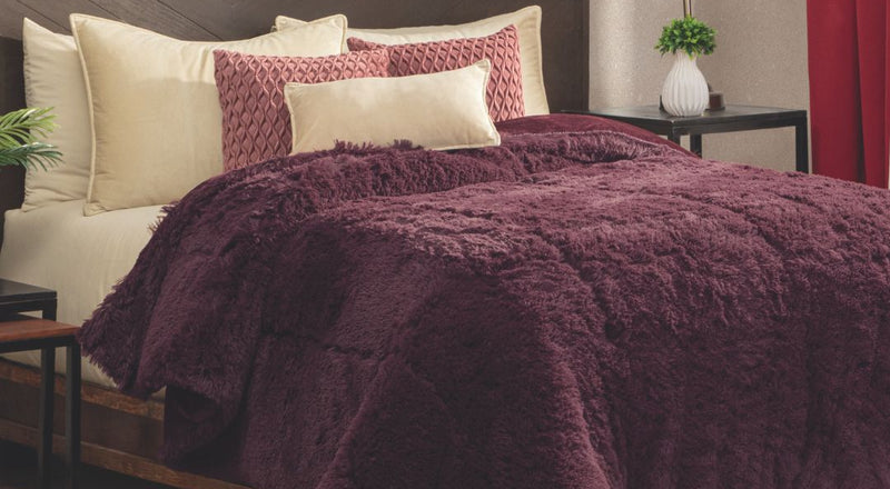 A bed featuring cream and burgundy pillows, a plush Cobertor Platinum Extra Suave Nevada comforter by Intima Hogar in burgundy, a dark wood headboard, bedside tables with lamps and plants, and a red curtain next to a beige wall.