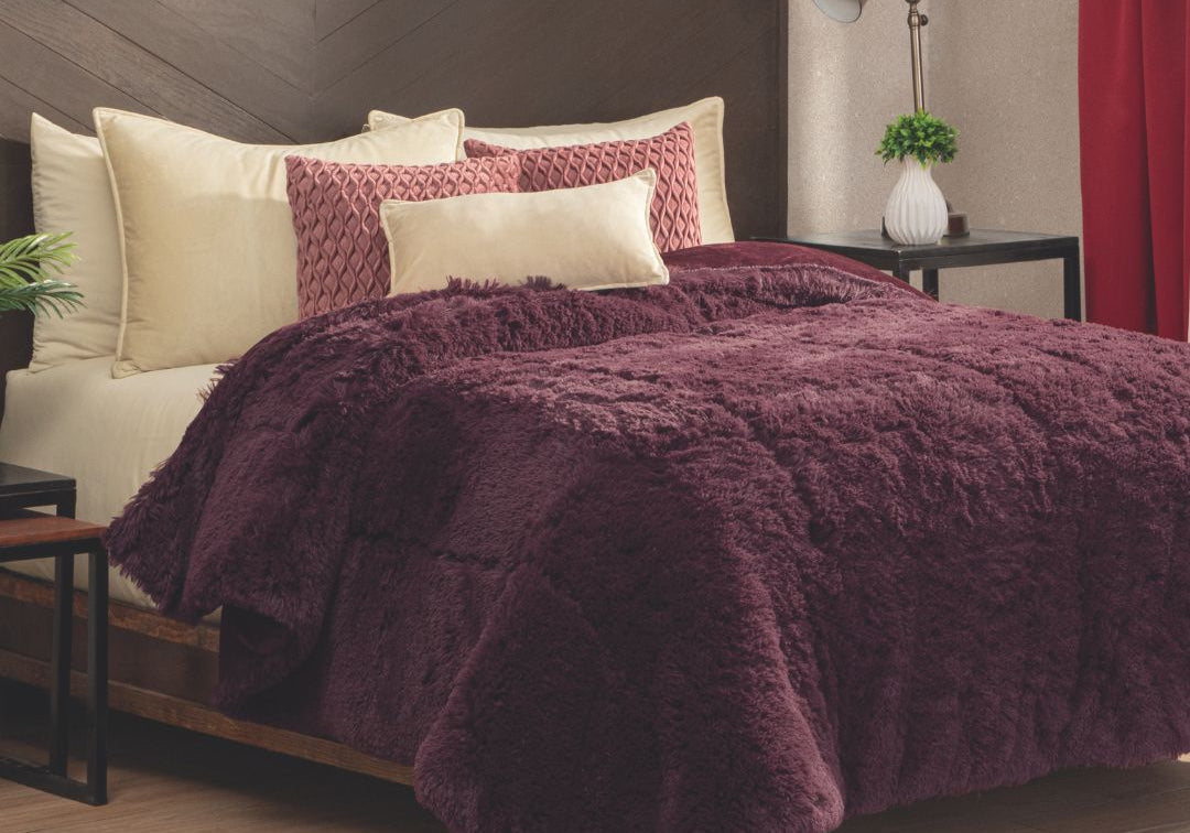 A bed featuring cream and burgundy pillows, a plush Cobertor Platinum Extra Suave Nevada comforter by Intima Hogar in burgundy, a dark wood headboard, bedside tables with lamps and plants, and a red curtain next to a beige wall.