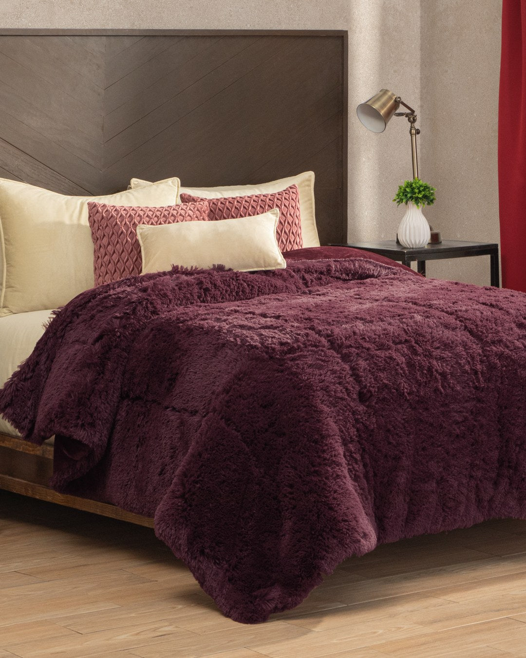 A neatly made bed with the ultra-soft Cobertor Platinum Extra Suave Nevada by Intima Hogar in a rich burgundy shade, paired with cream and burgundy accent pillows, a wooden headboard, a small lamp, a potted plant on the nightstand, and red curtains creates a cozy bedroom filled with calidez and textura extra suave for ultimate relaxation.