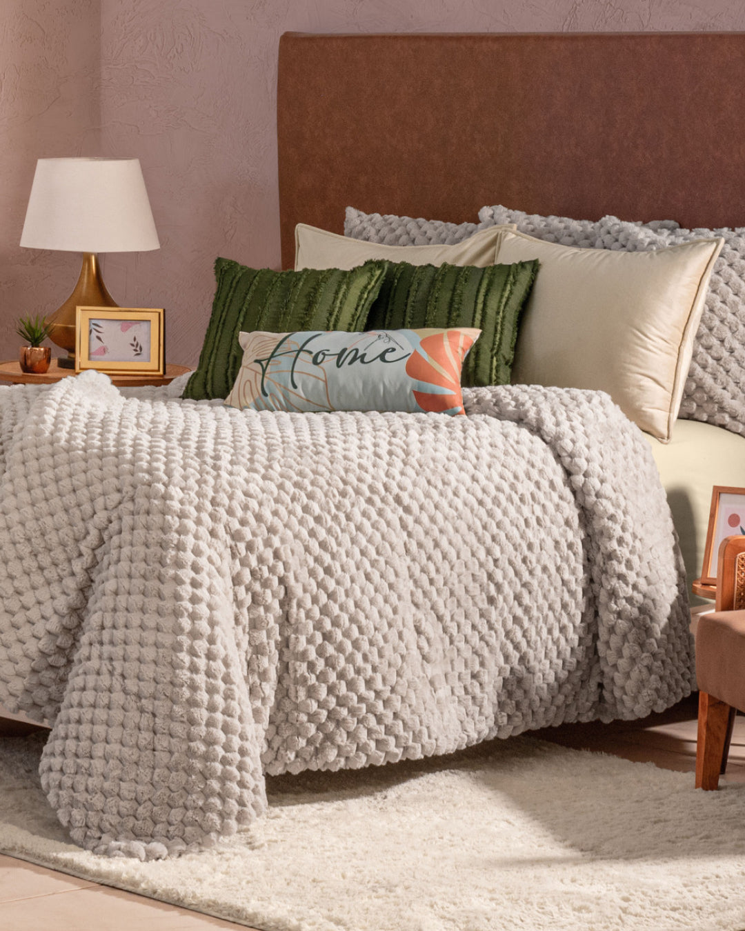 A cozy bed featuring the Cobertor Platinum Extra Suave Nickel by Intima Hogar, textured in a soft gray, is complemented by green and decorative pillows, a lamp, and framed photos on nightstands—all set in a softly lit bedroom that radiates calidez suave y tono neutro.