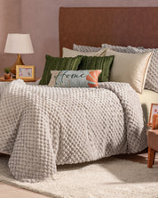 A cozy bed featuring the Cobertor Platinum Extra Suave Nickel by Intima Hogar, textured in a soft gray, is complemented by green and decorative pillows, a lamp, and framed photos on nightstands—all set in a softly lit bedroom that radiates calidez suave y tono neutro.