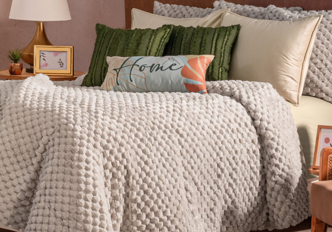 A cozy bed featuring the Cobertor Platinum Extra Suave Nickel by Intima Hogar, textured in a soft gray, is complemented by green and decorative pillows, a lamp, and framed photos on nightstands—all set in a softly lit bedroom that radiates calidez suave y tono neutro.