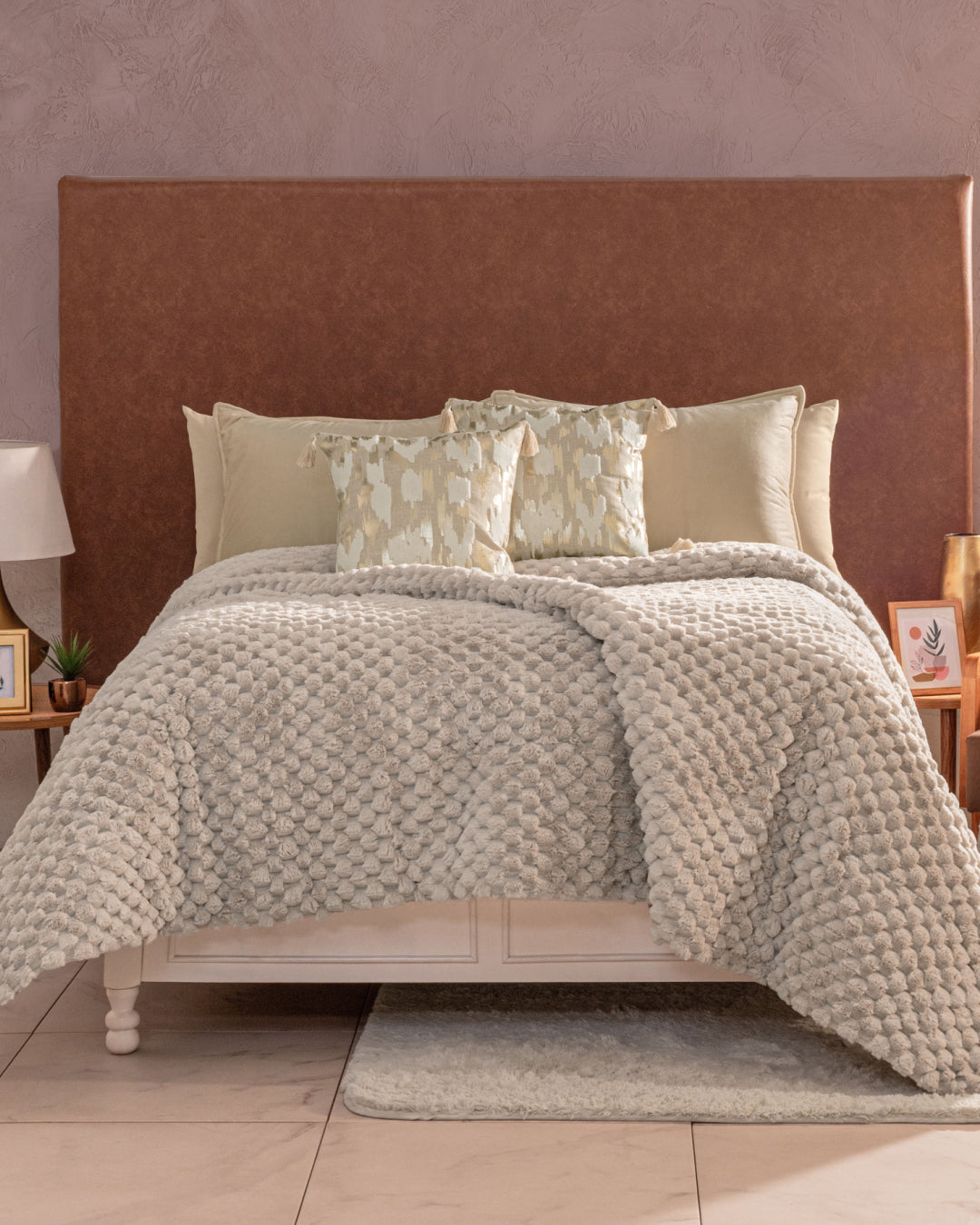 A neatly made bed is dressed with the Cobertor Platinum Extra Suave Nickel from Intima Hogar, a textured beige comforter featuring relieve afelpado, coordinated pillows, and a large brown headboard. The room’s taupe walls, wooden nightstands, lamps, and framed photos on either side create an atmosphere of calidez suave in a tono neutro.