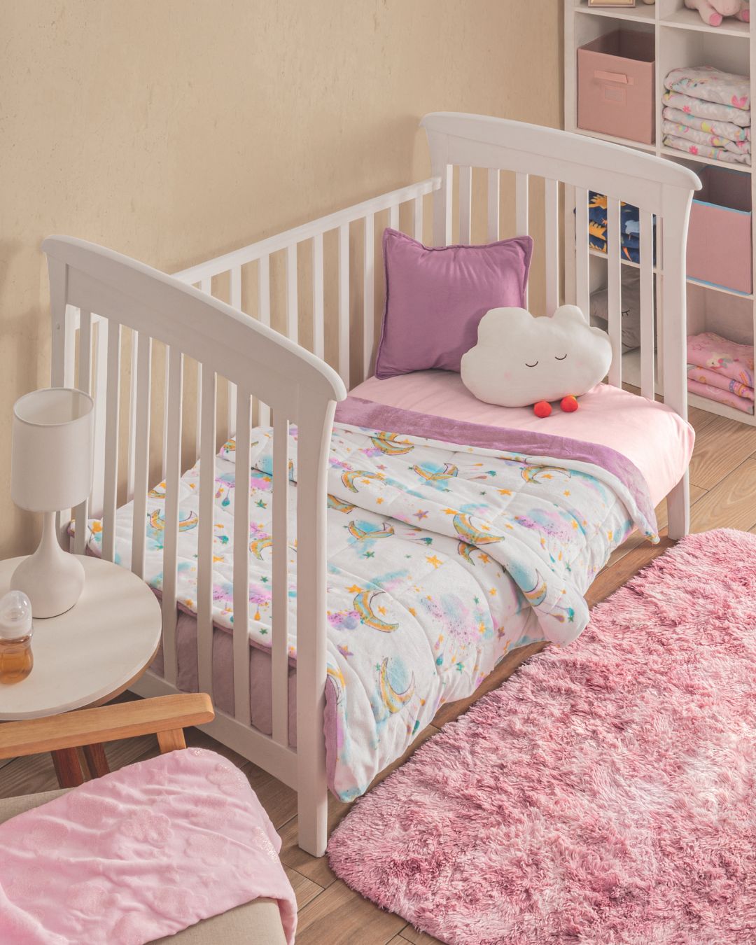 The Cobertor Flannel Baby Nube de Intima Hogar, junto a una cama con barandales blancos, ropa de cama en tonos pastel y cojín en forma de nube, crea un ambiente acogedor. Alfombra rosa y estantes con toallas completan el cuarto.