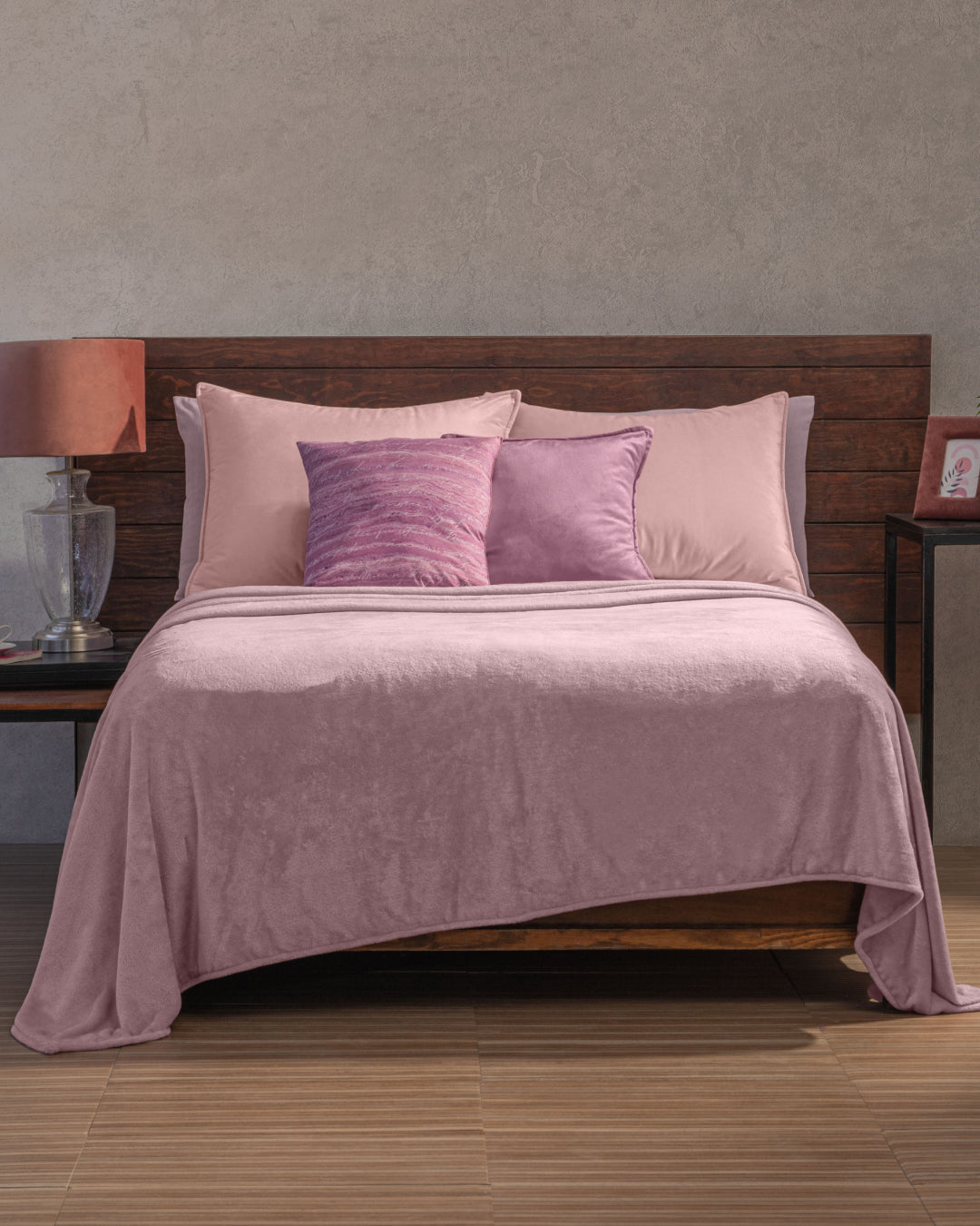 A bed with a wooden headboard is dressed in pink bedding and the Cobertor Flannel Ligero Ontario Lila by Intima Hogar, complemented by matching pillows. One nightstand holds a lamp and glass decor, while the other displays a framed photo. The room features neutral walls and a wood floor.