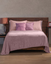 A bed with a wooden headboard is dressed in pink bedding and the Cobertor Flannel Ligero Ontario Lila by Intima Hogar, complemented by matching pillows. One nightstand holds a lamp and glass decor, while the other displays a framed photo. The room features neutral walls and a wood floor.