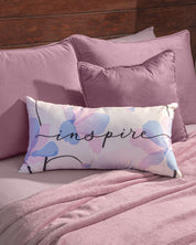A decorative rectangular pillow with pastel floral patterns and the word “inspire” in script lies on a mauve bed with matching square pillows, sheets, and the Cobertor Flannel Ligero Ontario Lila by Intima Hogar, adding an extra touch of warmth and coziness.