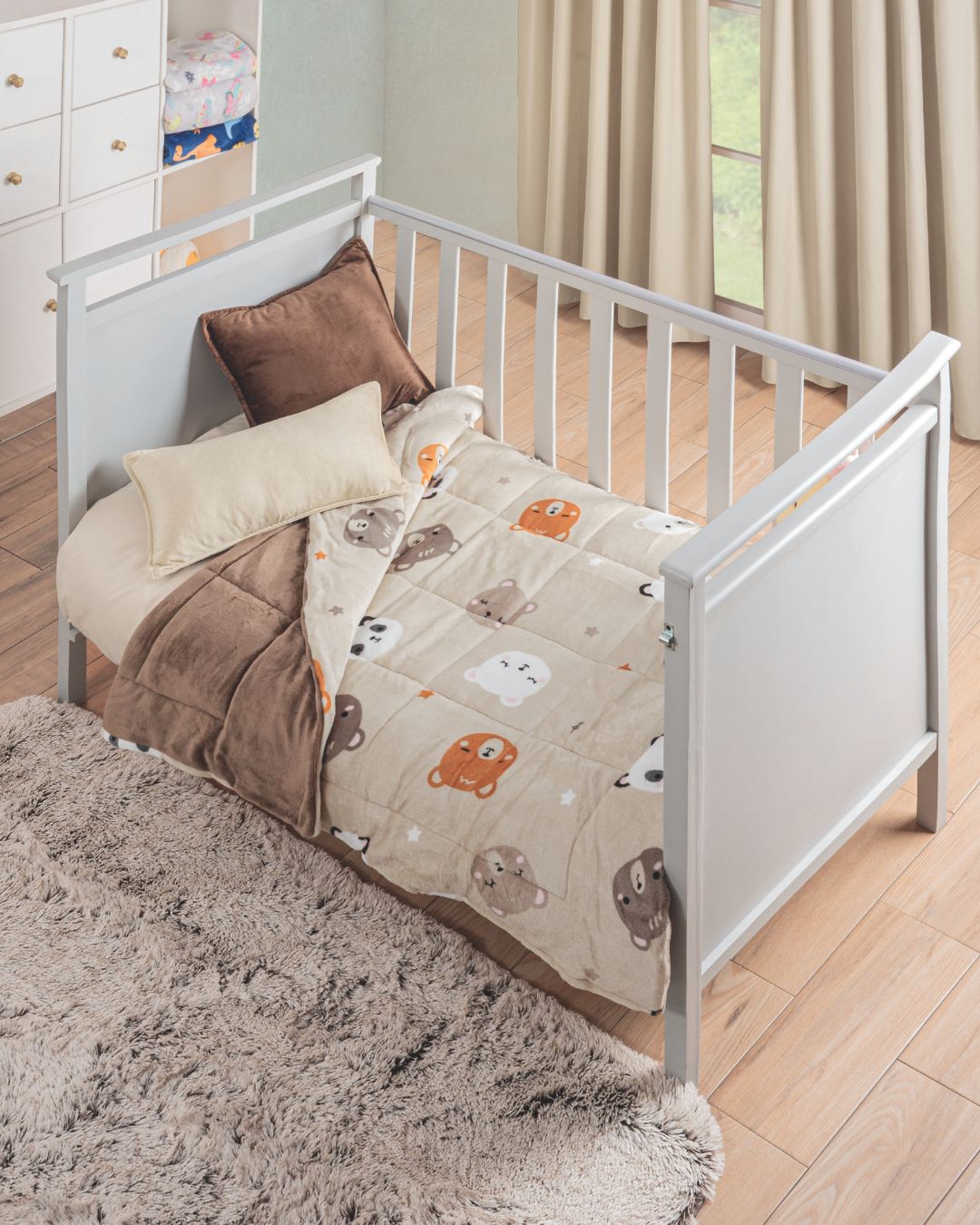 A white crib with the Cobertor Flannel Baby Ositos by Intima Hogar, featuring adorable bear faces in brown and cream, rests on a wooden floor with a fluffy gray rug, creating an inviting space softly lit by sheer curtains.