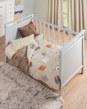 A white crib with the Cobertor Flannel Baby Ositos by Intima Hogar, featuring adorable bear faces in brown and cream, rests on a wooden floor with a fluffy gray rug, creating an inviting space softly lit by sheer curtains.