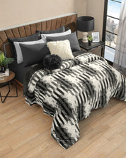 A modern bedroom featuring the Cobertor Flannel Extra Suave Oslo by Intima Hogar, adorned with black and gray pillows, a fluffy white pillow, a sleek black lamp, and a round side table. Sunlight streams through a large window, highlighting the geometric black-and-white patterns of the comforter and capturing the essence of diseño nórdico.
