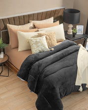 A neatly made bed with beige and cream pillows, a textured throw pillow, and the Cobertor Flannel Extra Suave Oslo de Intima Hogar—a plush dark gray comforter featuring patrones geométricos—layered with a white throw. Beside the bed sits a table holding a lamp, photo frame, and small plant, all set in a cozy bedroom inspired by diseño nórdico.
