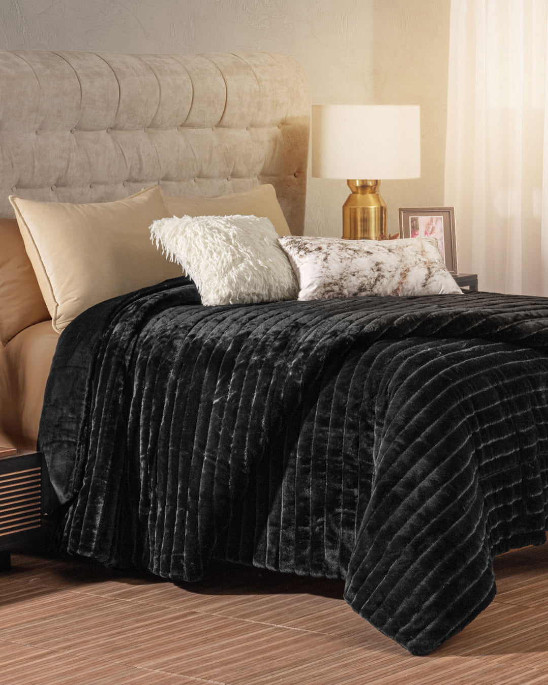 A neatly made bed is styled with the Intima Hogar Cobertor Platinum Extra Suave Petróleo—a luxuriously soft, black quilted blanket featuring líneas afelpadas—paired with beige sheets and three decorative pillows. Next to the upholstered headboard, a gold lamp, photo frame, and small decor items are arranged on the bedside table.