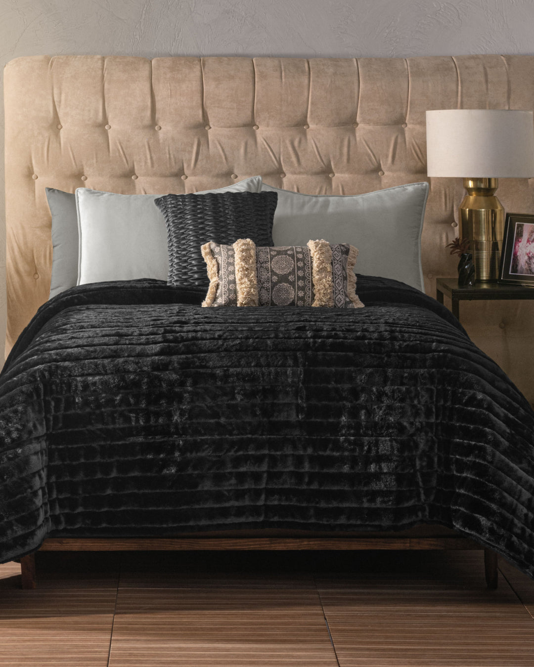 A bed with a beige tufted headboard, gray and patterned pillows, and the Cobertor Platinum Extra Suave Petróleo by Intima Hogar—featuring cobertura abrigadora—rests beneath a lamp and photo frame atop a side table, all set against a textured wall.