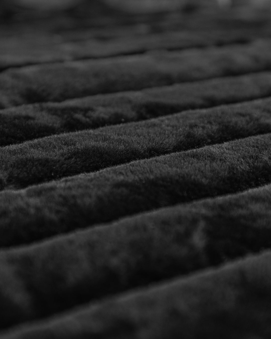 Close-up of the Cobertor Platinum Extra Suave Petróleo by Intima Hogar in black, showcasing its soft, velvety surface with parallel horizontal ridges. The lighting accentuates the plush, fuzzy fabric and highlights the depth between the líneas afelpadas, emphasizing its irresistible textura acanalada.