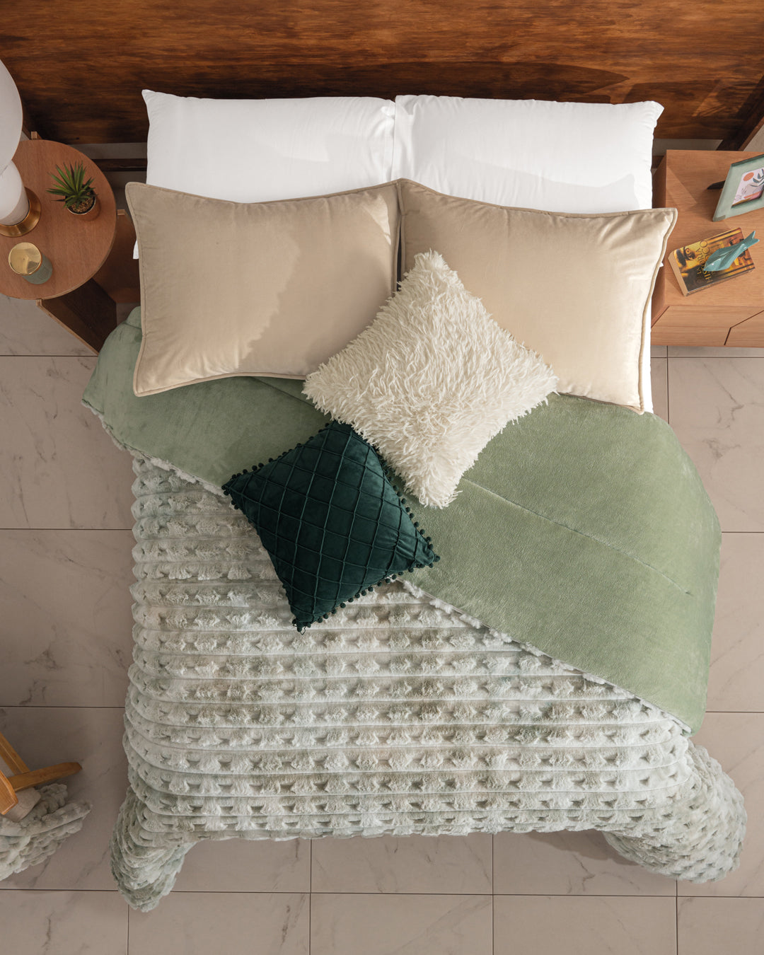 Overhead view of a bed with light green bedding, Intima Hogar’s Cobertor Platinum Extra Suave Siberia, a textured blanket, beige pillows, and white and dark green decorative cushions beside wooden nightstands on a tiled floor.