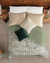 Overhead view of a bed with light green bedding, Intima Hogar’s Cobertor Platinum Extra Suave Siberia, a textured blanket, beige pillows, and white and dark green decorative cushions beside wooden nightstands on a tiled floor.