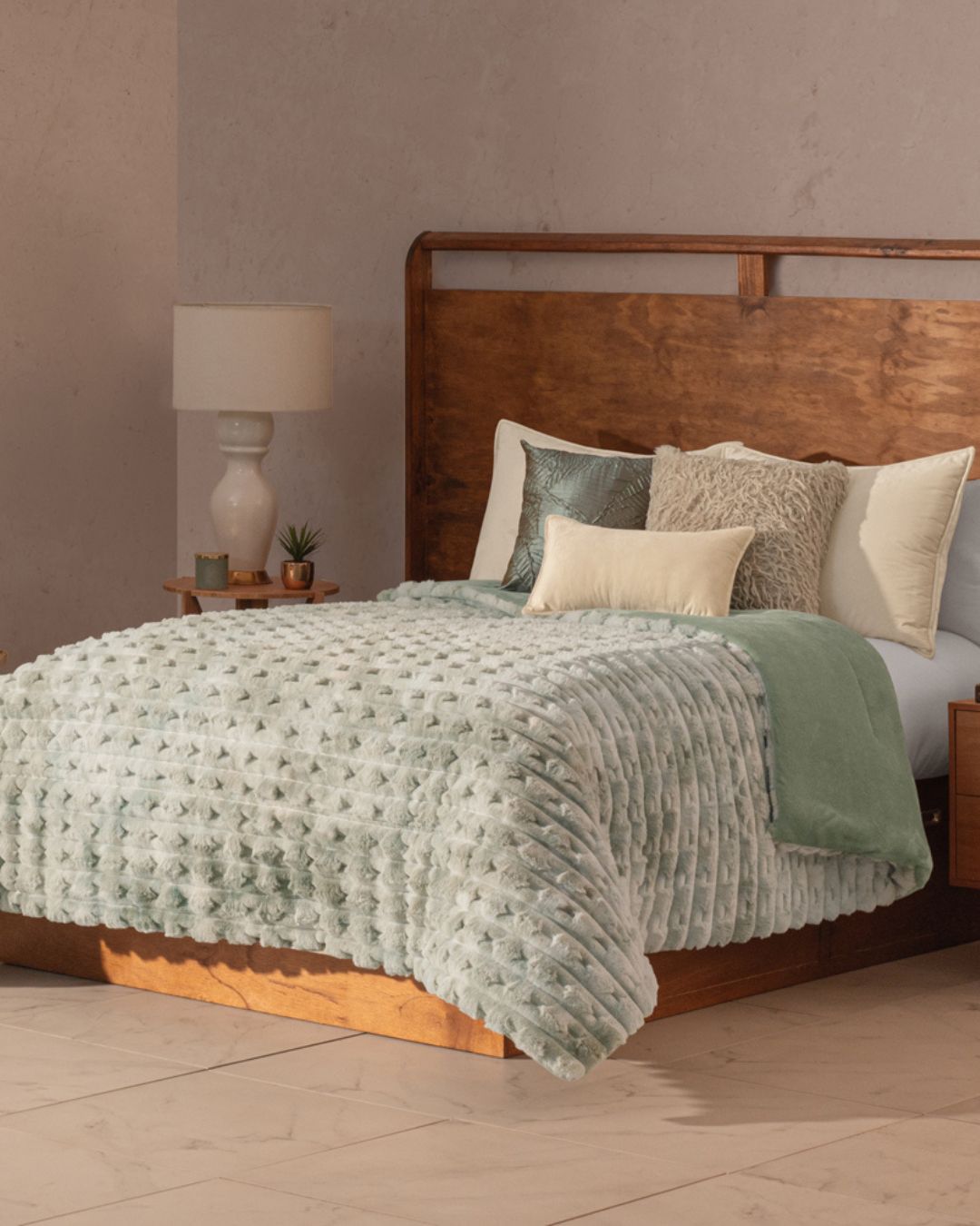 A cozy bedroom features the Intima Hogar Cobertor Platinum Extra Suave Siberia on a wooden bed frame, with assorted pillows, a wooden nightstand, lamp, books, and small plants against light walls and tiled floors.