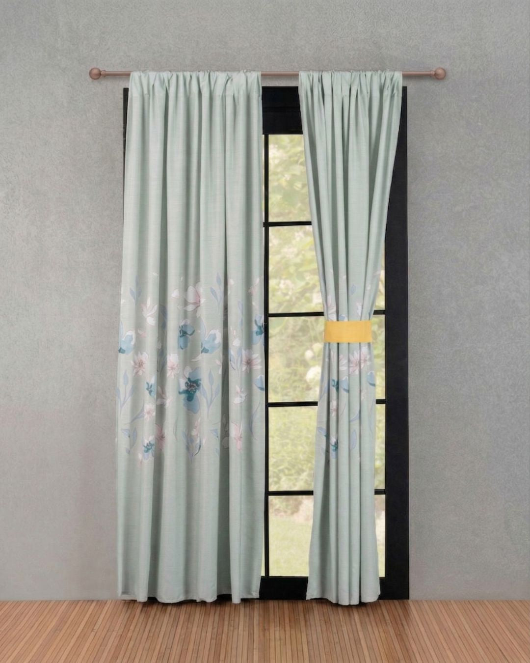 El Juego de Cortinas Celina de Intima Hogar en verde claro con suaves flores viste la ventana; una cortina está recogida con una banda amarilla, la otra cuelga recta, creando armonía y mostrando la vista exterior.