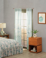 A cozy bedroom features light blue floral bedding and the Juego de Cortinas Celina by Intima Hogar. A wooden nightstand with a lamp, clock, and plant stands by the bed, sunlight pours in, and abstract art adorns the gray wall for un estilo armonioso.