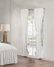 Las cortinas blancas Juego de Cortinas Paloma de Intima Hogar, recogidas en una puerta de vidrio, dejan entrar la luz a una habitación con cama, ropa blanca, lámpara dorada y piso de madera; se ve un patio con plantas afuera.