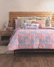 A cozy bed with a wooden headboard features the Intima Hogar Edredón Súper Suave Bora Bora, colorful leaf patterns, and multiple pillows (one says "inspire"). The bedside table holds a lamp, vase, photo frame, books, and decor items.