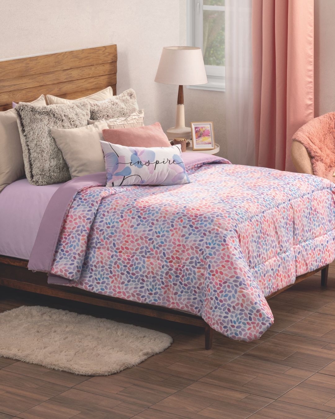 A cozy bedroom with a wooden bed, pastel floral bedding, the Edredón Súper Suave Bora Bora by Intima Hogar in vibrant tones, various pillows including one that says “Inspire,” a bedside lamp, framed photo, pink curtains, and a soft white rug.