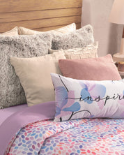 A neatly made bed features fluffy cream and gray pillows, a pink cushion, a pastel floral “inspire” pillow, and is dressed with an Intima Hogar Edredón Súper Suave Bora Bora in lavender and dotted pastels.