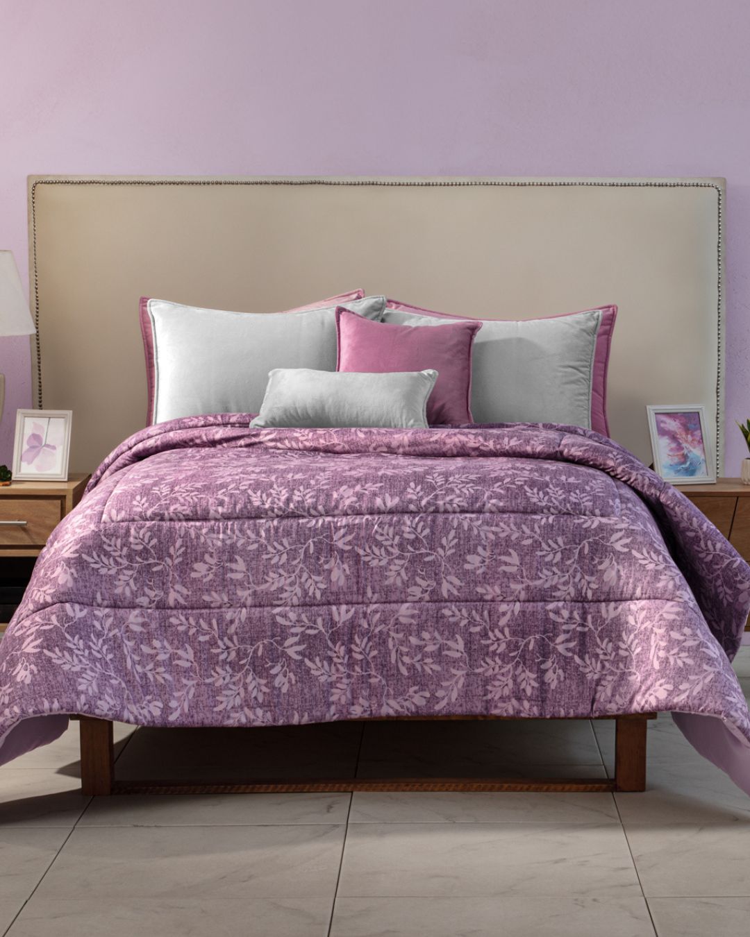 A neatly made cama with a light beige headboard is dressed in the Edredón Súper Suave Maya by Intima Hogar, featuring purple florals and accented by white, pink, and purple pillows. Wooden nightstands with lamps and framed art complete the look.