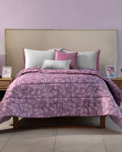 A neatly made cama with a light beige headboard is dressed in the Edredón Súper Suave Maya by Intima Hogar, featuring purple florals and accented by white, pink, and purple pillows. Wooden nightstands with lamps and framed art complete the look.
