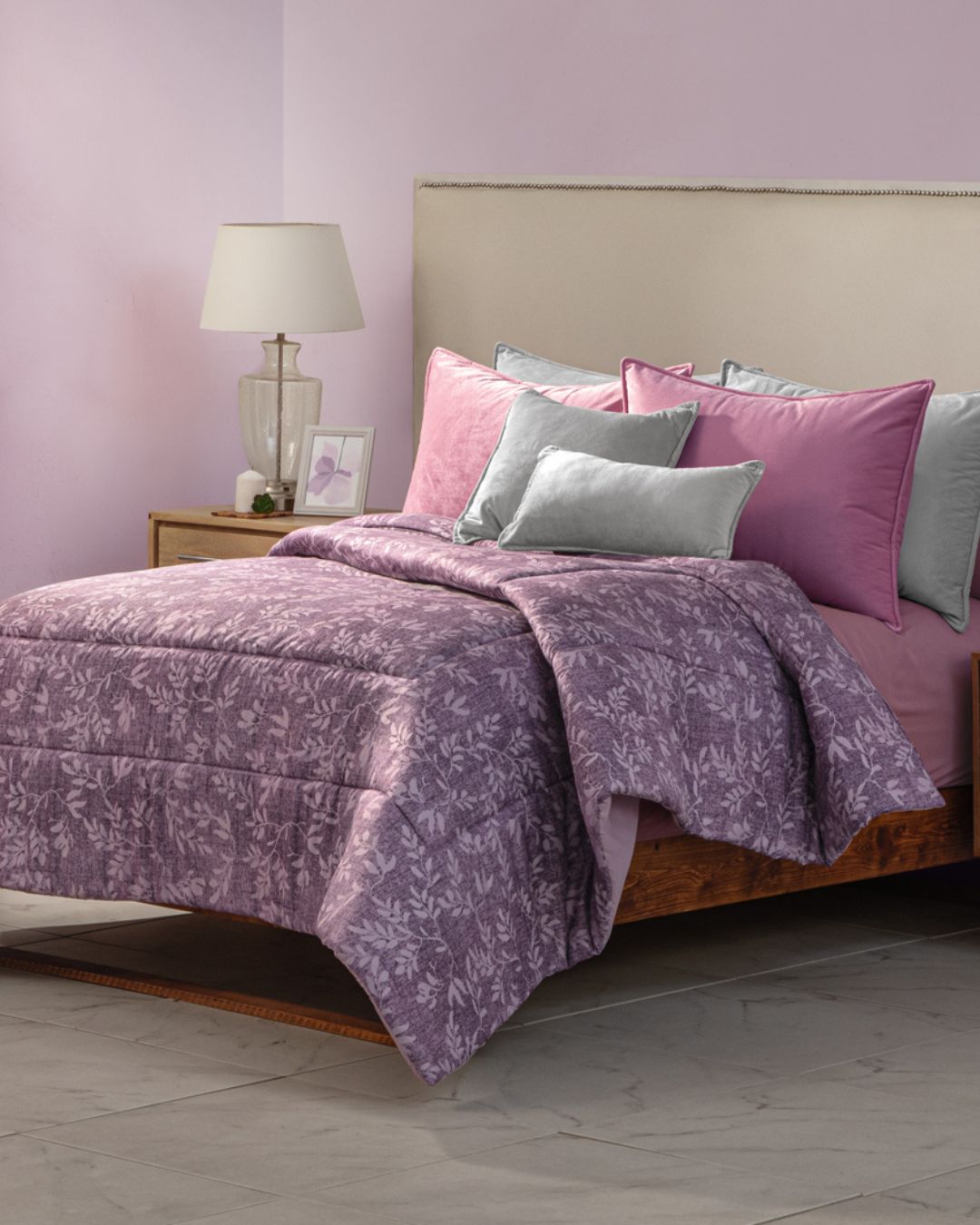 A neatly made bed features the Intima Hogar Edredón Súper Suave Maya, with pink and mauve floral patterns, matching pillows, a light beige headboard, a wooden nightstand with a lamp and photo frame, all set against a light purple wall.