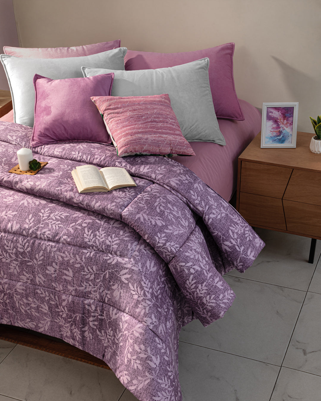 A cozy bed features the Edredón Súper Suave Maya by Intima Hogar in a purple floral design, paired with lavender and gray pillows. An open book and cup rest on a plate, while a wooden nightstand holds decor atop a tiled floor.