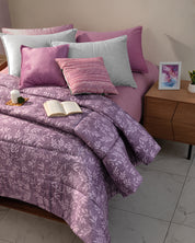 A cozy bed features the Edredón Súper Suave Maya by Intima Hogar in a purple floral design, paired with lavender and gray pillows. An open book and cup rest on a plate, while a wooden nightstand holds decor atop a tiled floor.
