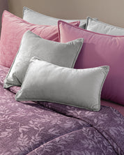 The bed showcases the Edredón Súper Suave Maya by Intima Hogar, styled with two gray velvet pillows and three pink pillows. Its purple floral-patterned design creates a soft, cozy, and elegant atmosphere in the room.