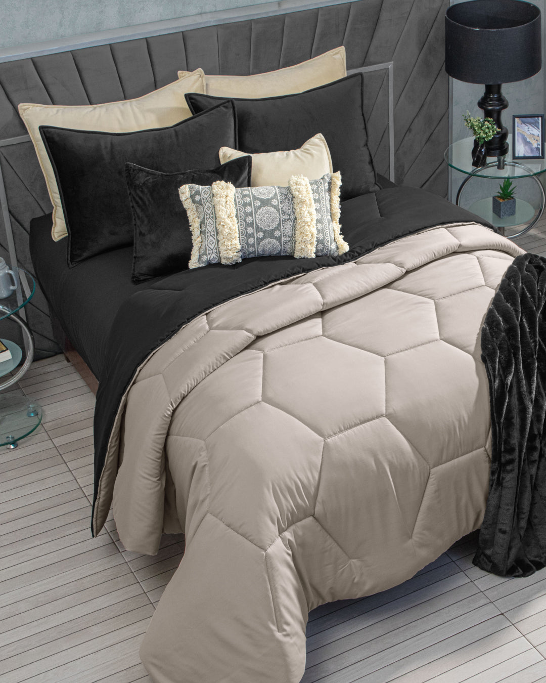 A neatly made recámara featuring the Edredón Down Silky Oaxaca by Intima Hogar in beige and black, paired with decorative pillows, a black lamp, glass side tables, and a modern grey padded headboard creates a contemporary bedroom with cobertura visual and an estilo limpio.