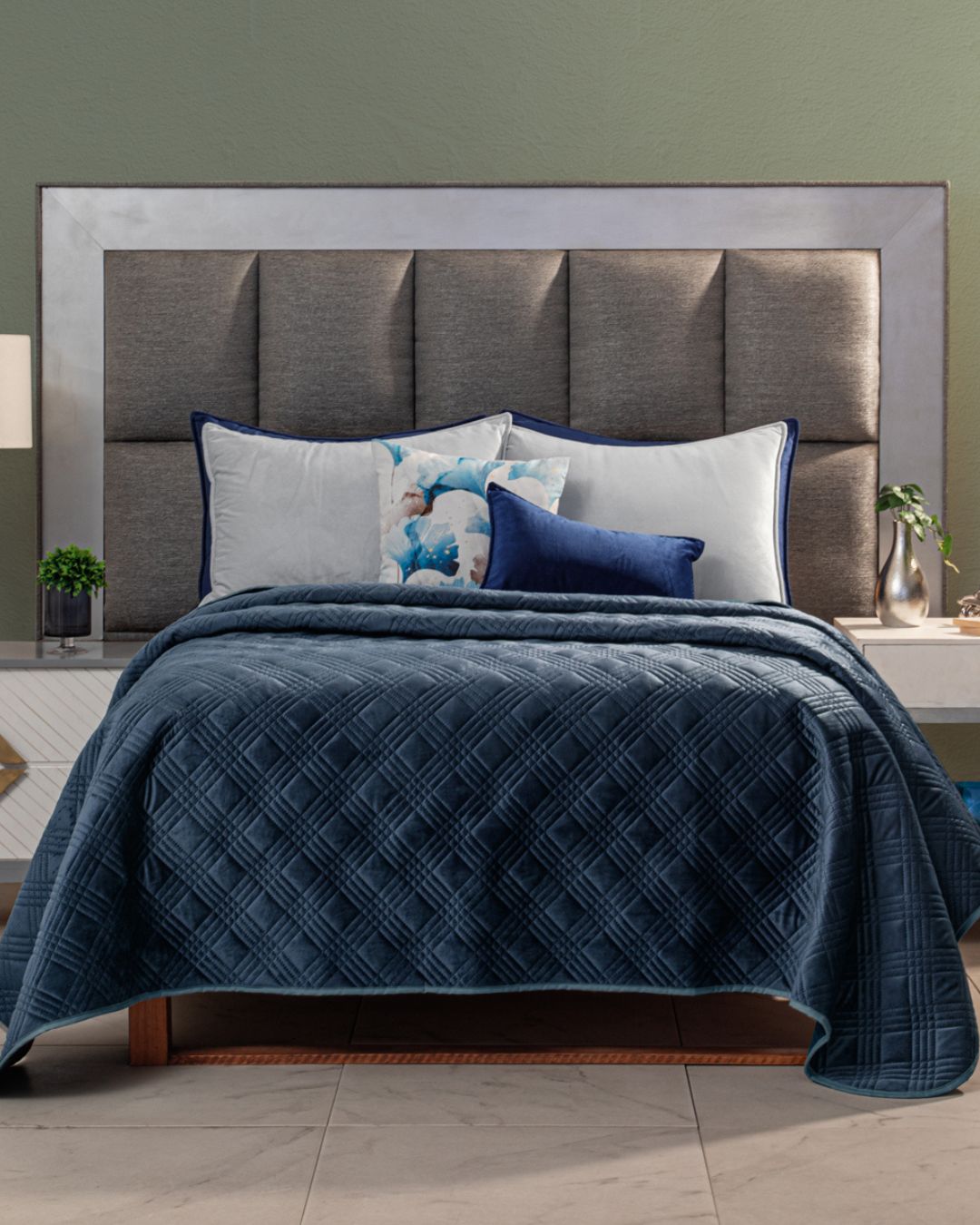 A neatly made bed with a tall, padded gray headboard features the Edredón Velvet Hielo by Intima Hogar, paired with white and blue pillows. Nightstands holding small plants and lamps rest on each side against a muted green wall.