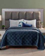 A neatly made bed with a tall, padded gray headboard features the Edredón Velvet Hielo by Intima Hogar, paired with white and blue pillows. Nightstands holding small plants and lamps rest on each side against a muted green wall.