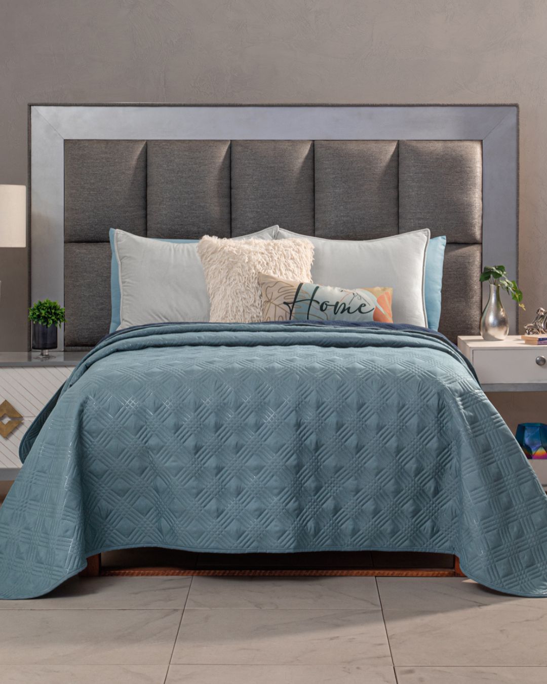 A bed with a padded gray headboard, white and blue pillows, a decorative Home pillow, and the Edredón Velvet Hielo by Intima Hogar in blue. Side tables with small plants and decor flank the setup.