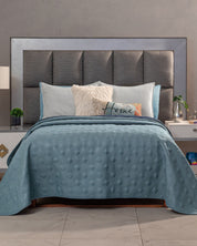 A bed with a padded gray headboard, white and blue pillows, a decorative Home pillow, and the Edredón Velvet Hielo by Intima Hogar in blue. Side tables with small plants and decor flank the setup.
