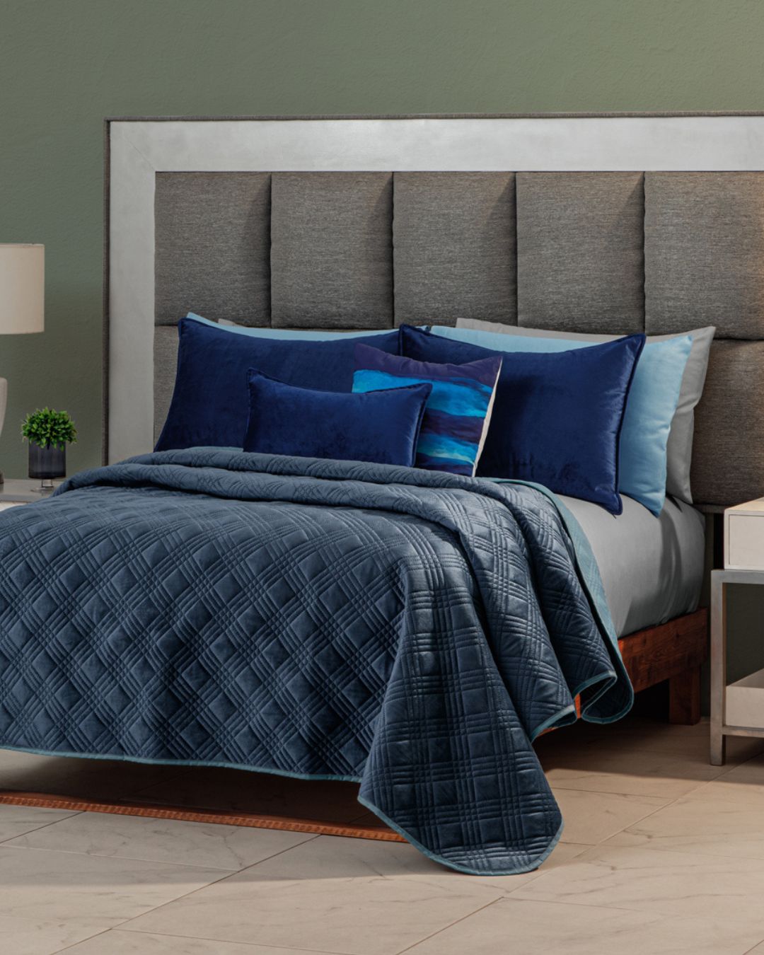 A neatly made bed features the Edredón Velvet Hielo by Intima Hogar, several blue and navy pillows, and a large padded headboard, set against a green wall with a white nightstand and table lamp nearby.
