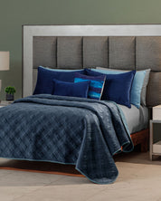 A neatly made bed features the Edredón Velvet Hielo by Intima Hogar, several blue and navy pillows, and a large padded headboard, set against a green wall with a white nightstand and table lamp nearby.