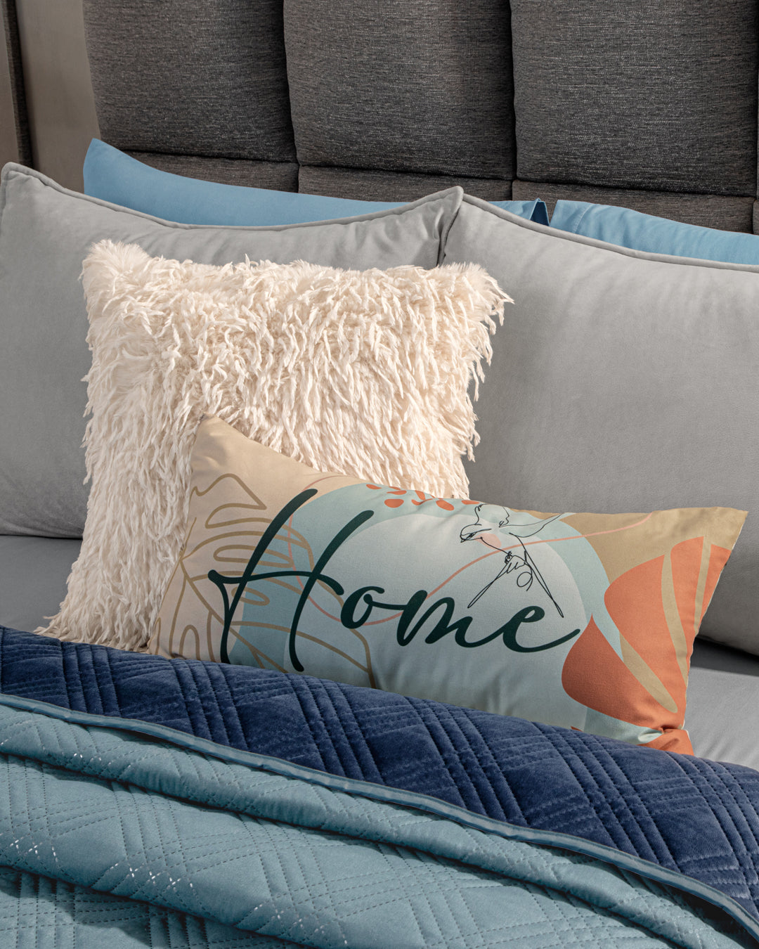 A bed dressed with the Edredón Velvet Hielo by Intima Hogar, blue and gray bedding, a fluffy cream pillow, and a decorative "Home" script pillow sits against a gray upholstered headboard.