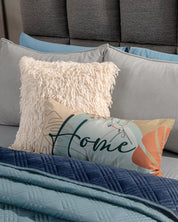 A bed dressed with the Edredón Velvet Hielo by Intima Hogar, blue and gray bedding, a fluffy cream pillow, and a decorative "Home" script pillow sits against a gray upholstered headboard.