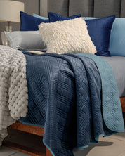A neatly made bed features the Edredón Velvet Hielo by Intima Hogar, in icy blue and light gray tones, paired with navy pillows, a cream textured cushion, a pale blue quilt, and a soft chunky gray knit blanket draped over the wooden frame.