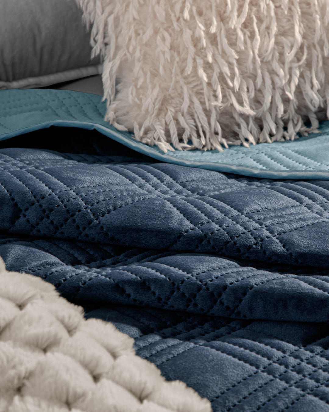 Close-up of layered bedding with Intima Hogar's Edredón Velvet Hielo, accented by a cream chunky knit blanket, light blue sheet, and a textured white pillow with shaggy tassels.