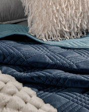 Close-up of layered bedding with Intima Hogar's Edredón Velvet Hielo, accented by a cream chunky knit blanket, light blue sheet, and a textured white pillow with shaggy tassels.