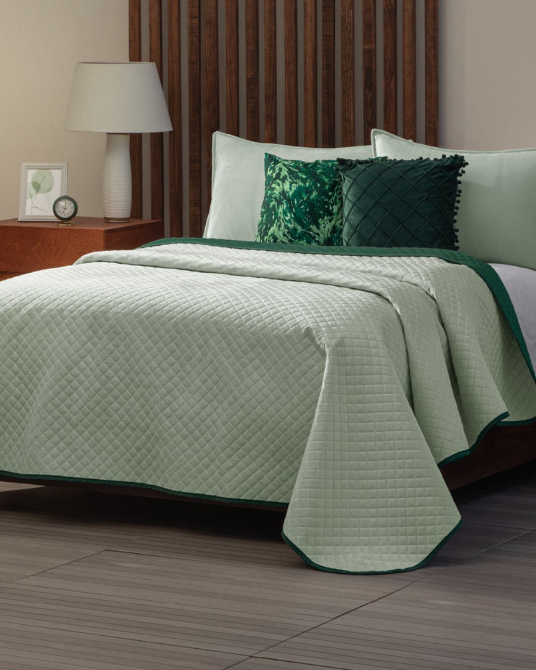 A neatly made bed features the Edredón Velvet Mundo by Intima Hogar, a reversible light green quilted bedding set with matching and decorative green pillows. A wooden headboard, lamp-lit nightstand, and framed art complete the natural ambiance.