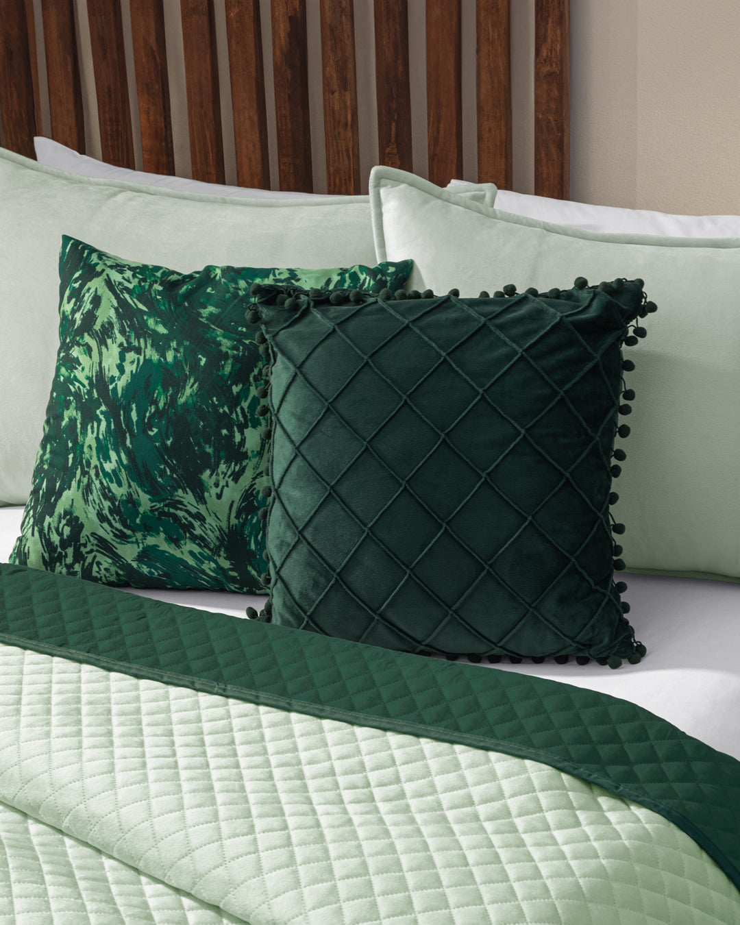 Two decorative pillows—a dark green quilted one with pom-pom trim and a green marbled one—sit on a bed dressed in the Edredón Velvet Mundo by Intima Hogar, with a wooden headboard in the background.
